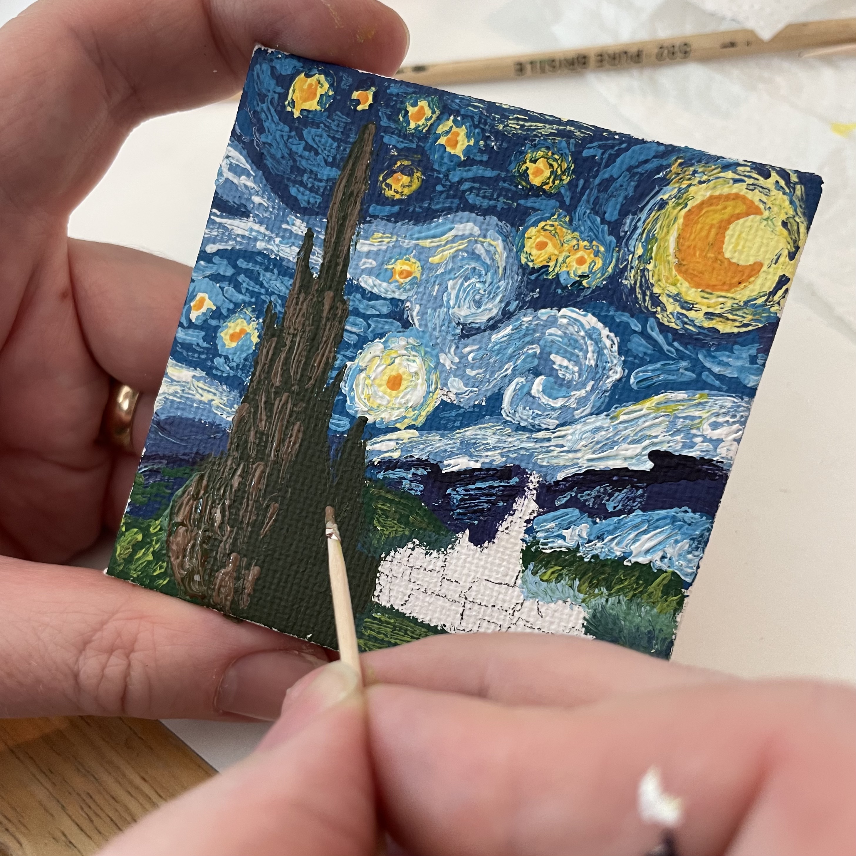 A close-up shows a person painting a miniature reproduction of Vincent van Gogh's The Starry Night. The small canvas is held in one hand while the other uses a toothpick to add brown details to the tall cypress tree in the foreground. Much of the swirling blue and white night sky, dotted with bright yellow stars and a crescent moon, is already complete, while part of the village below remains unpainted, revealing the white canvas. The artist's hands are in focus, with a wooden-handled brush and a paper towel visible in the softly blurred background.